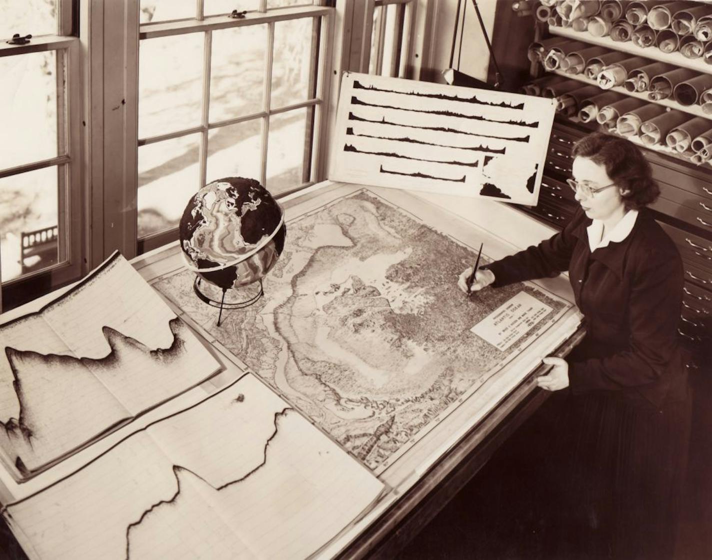 Marie Tharp poses in Lamont Hall with a physiographic diagram of the North Atlantic Ocean. Sounding records are visible on the left. A prototype of a globe she constructed with Bruce Heezen sits in the middle. Her profiles of the North Atlantic seabed are propped in the corner. Photo from the late 1950s.