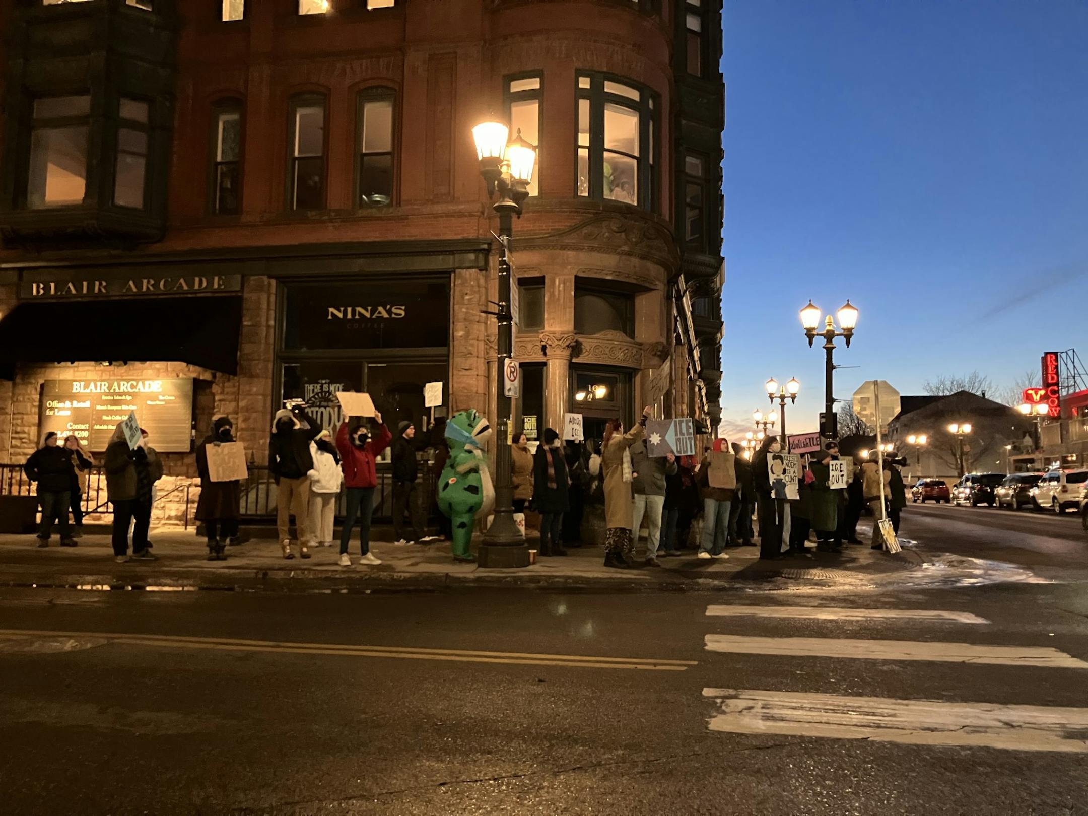Several people hold signs with slogans such as "ICE out" in front of an ornate brick building, and ne person wears an inflatable green frog costume. Street lamps glow and the sun has just set.