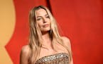 Margot Robbie in a gold strapless beaded gown in front of a red background.