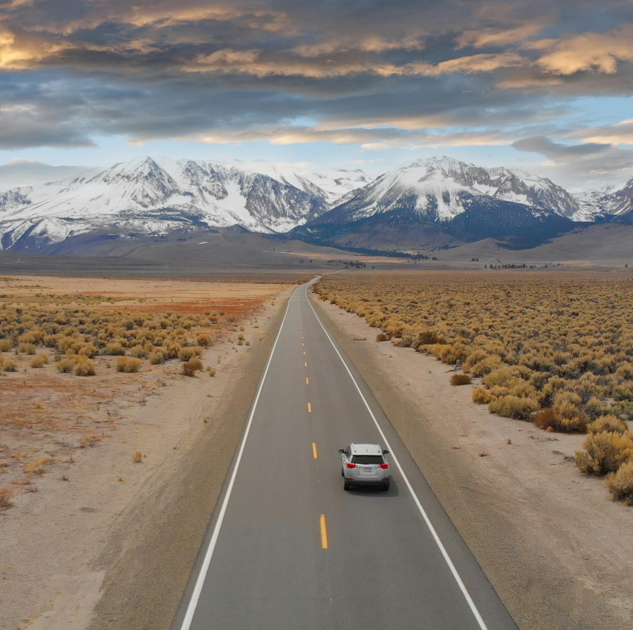 AERIAL: Large SUV drives down the empty highway leading to the spectacular Rockies. Flying above interstate running through the picturesque rugged countryside in Nevada. Breathtaking mountain range.
