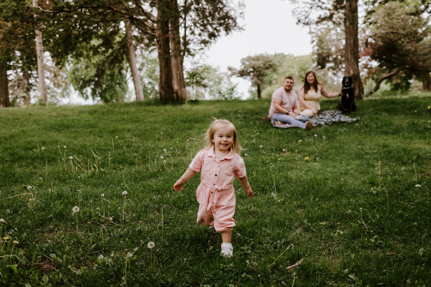 A smiling blond toddler in a pink jumpsuit sprints toward the camera in a park while a couple and a dog sit in the background on a picnic blanket.