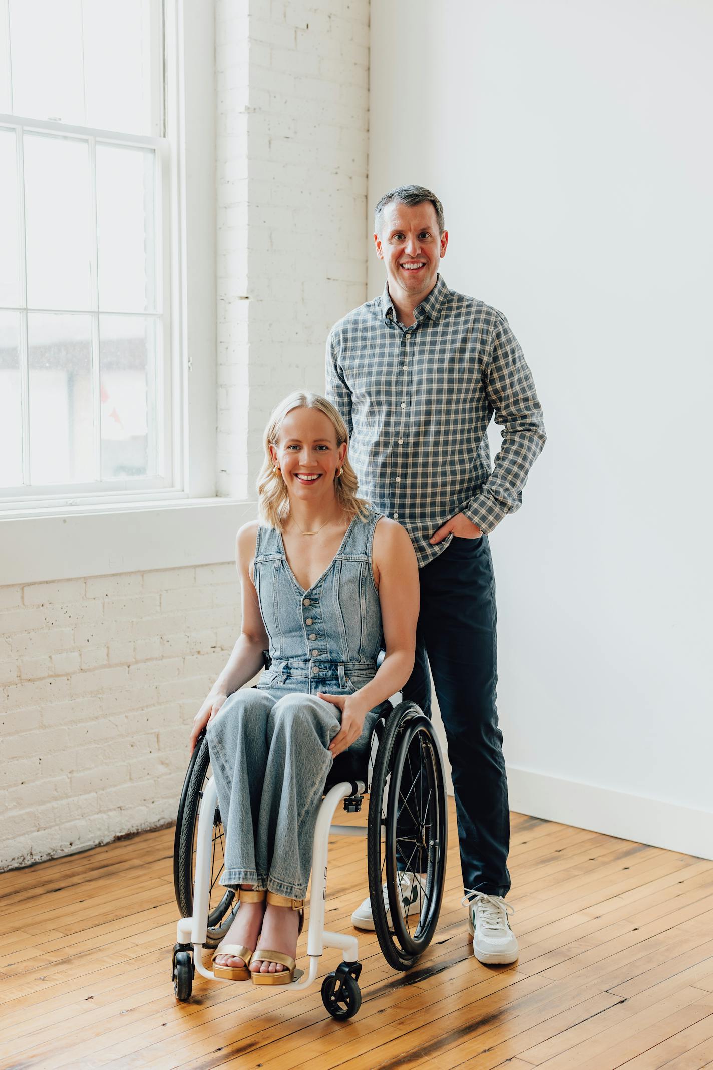 A woman in a wheelchair and a man standing smile toward the camera in a sunlit studio space with white walls and hardwood floors.