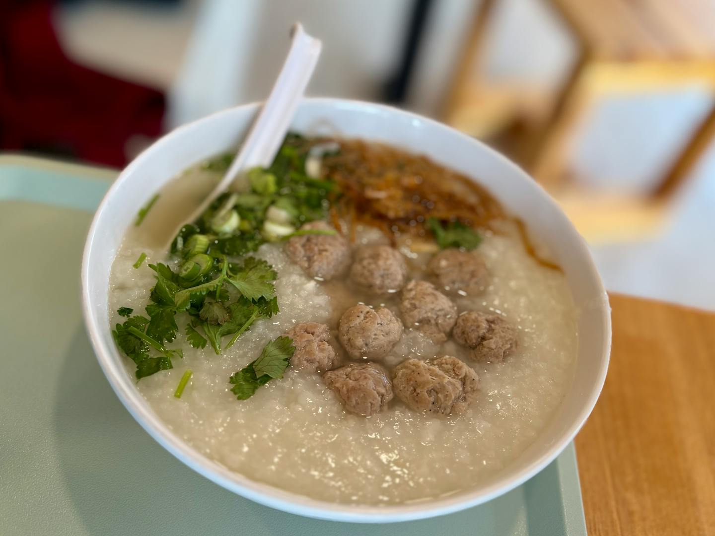 A large bowl of brothy chicken congee is topped with green onions, cilantro, fried shallots and small meatballs.