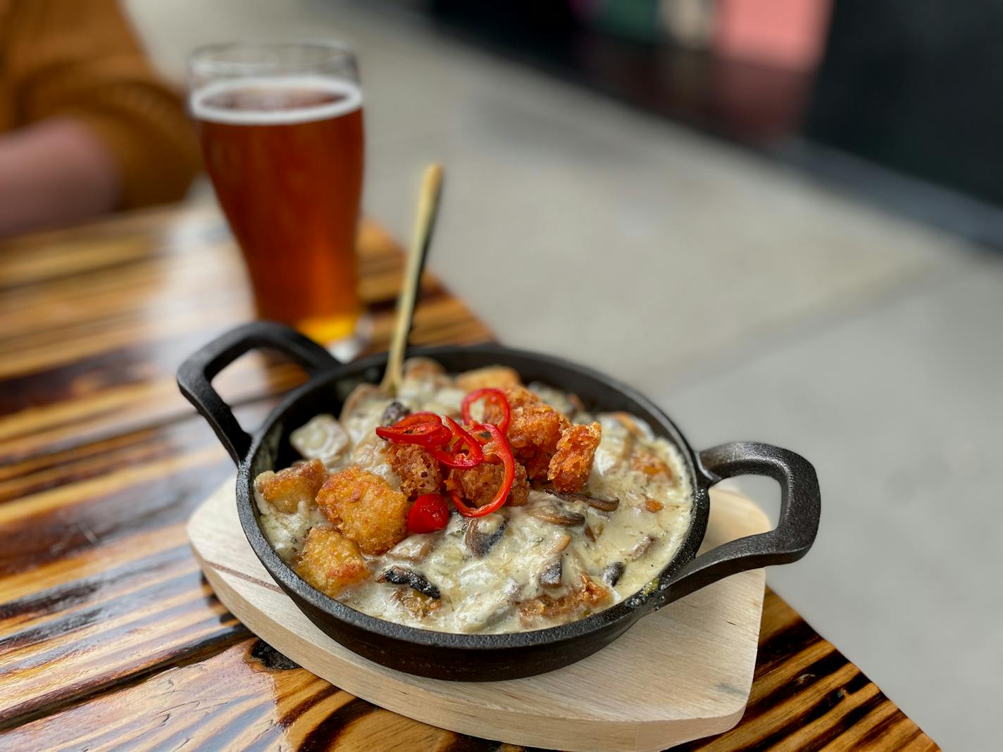 A cast iron pan filled with mushroom cream sauce, topped with tater tots and slices of a red chili on a wooden table outside, next to an amber colored beer.