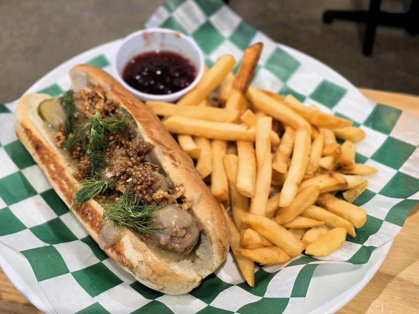 A sub sandwich with meatballs, whole grain mustard and green fronds of dill, on a plate with a pile of fries and a cup of red berry jam