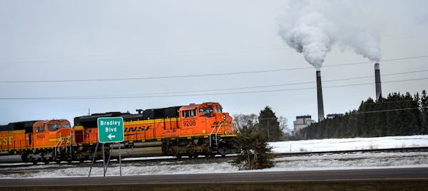 A BNSF Railway train waits on the tracks near Becker, Minn. with the twin smokestacks of Xcel Energy&#x2019;s coal-fired Sherco power plant emitting s