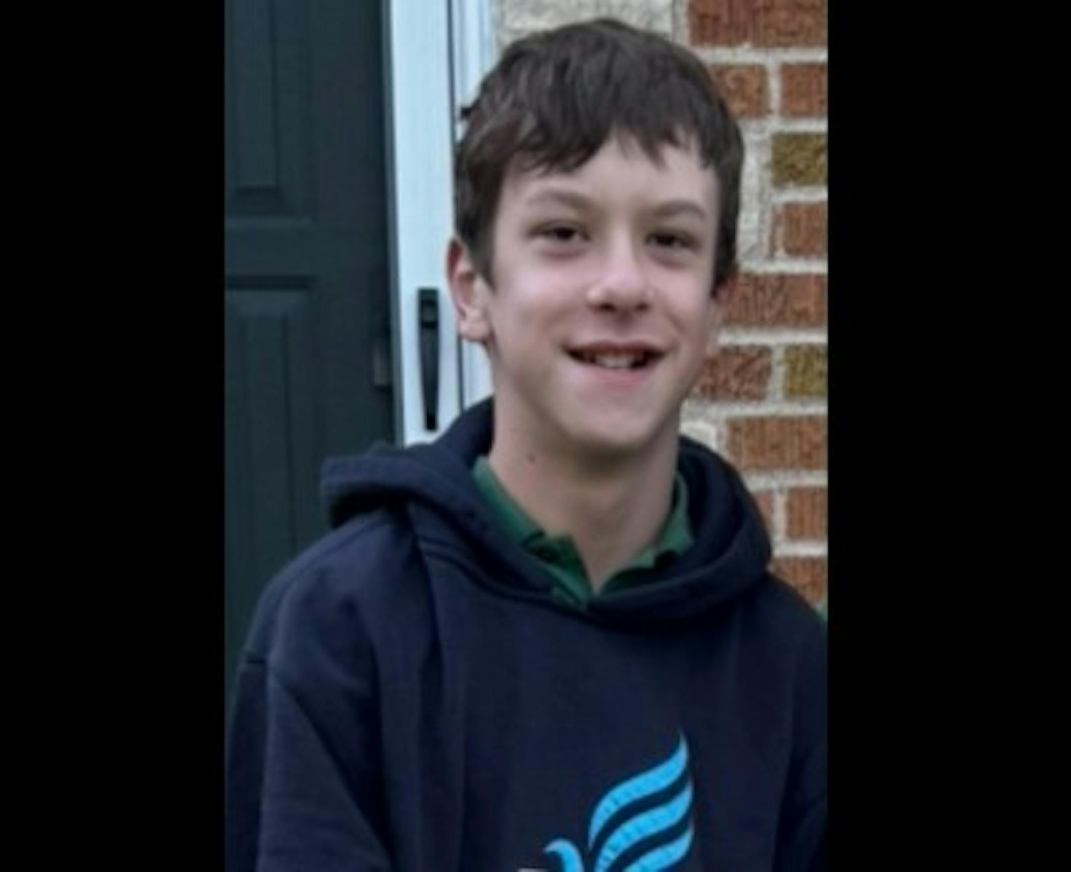 A boy in a dark hoodie smiles in front of a brick house.
