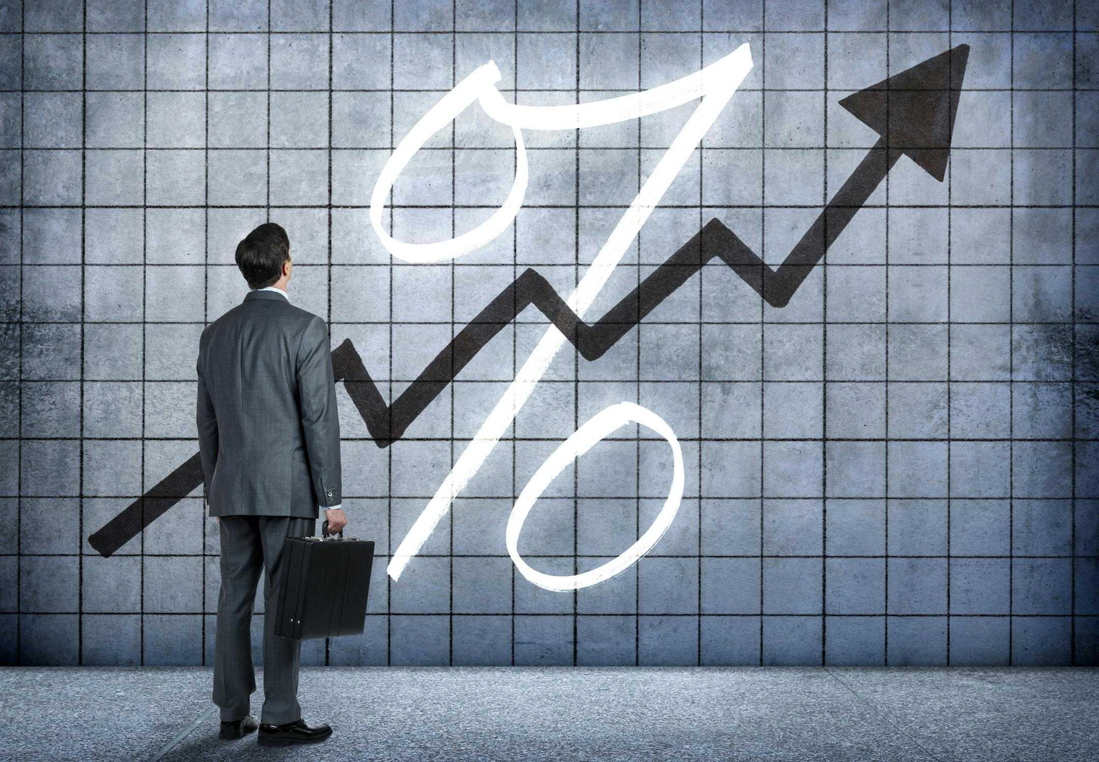 A businessman, standing while holding a briefcase, looks up at a chart on a wall that indicates interest rates are rising. istock