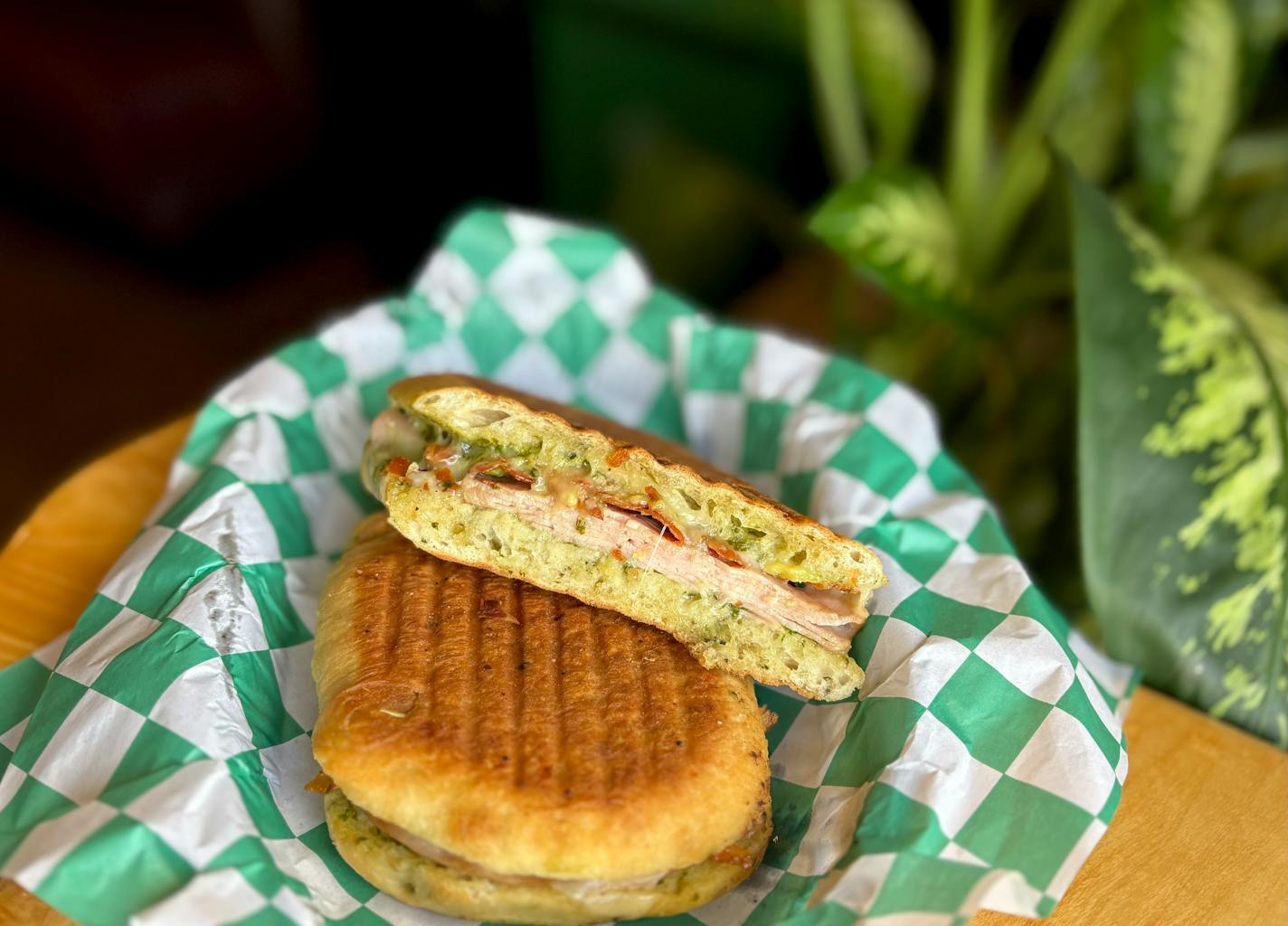 A turkey and bacon panini photographed in a basket with white and green wax paper. Background is a wooden table with a variegated green plant in the background.