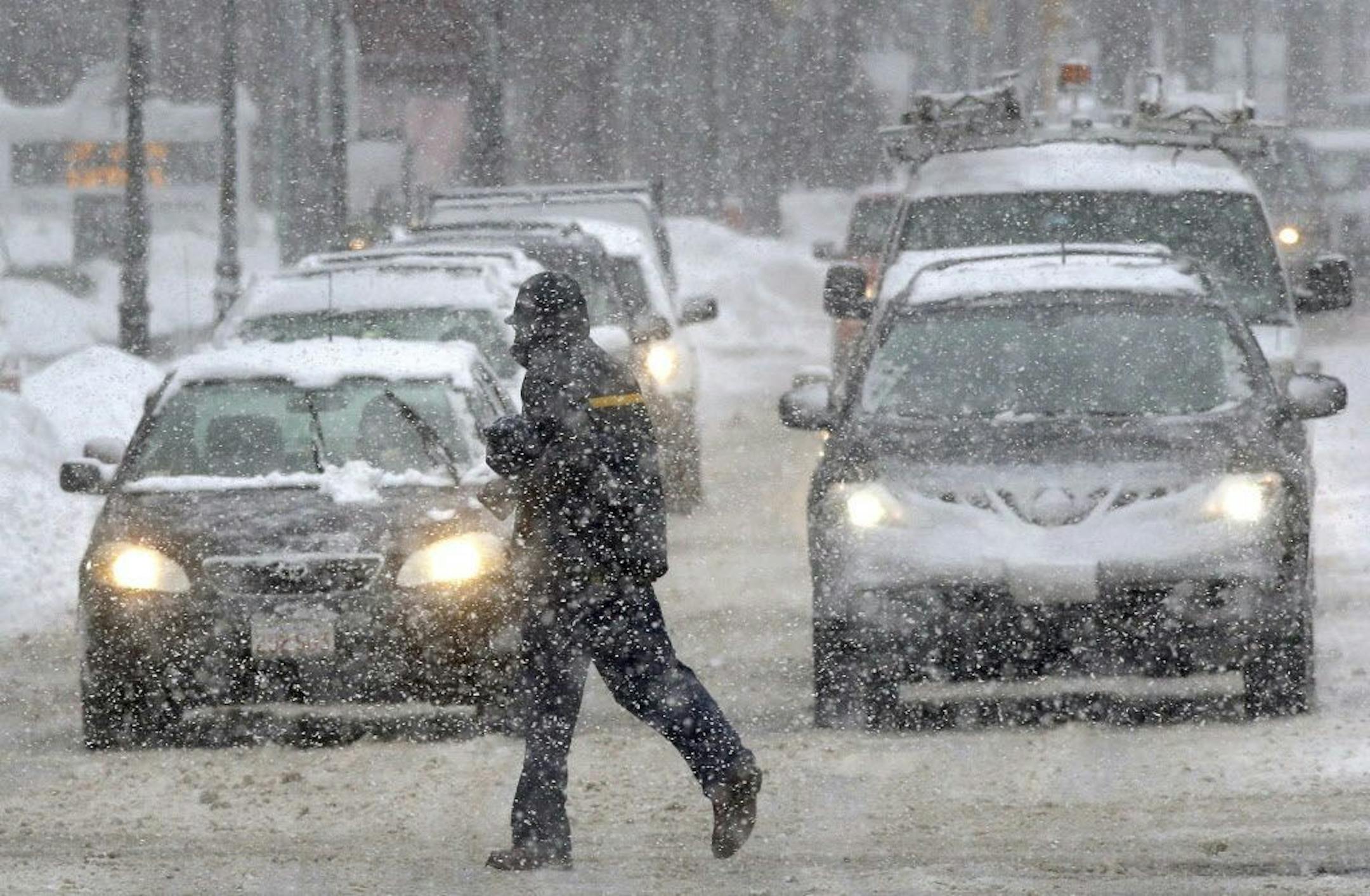 A pedestrian crosses the street in front of vehicles during a snowstorm.