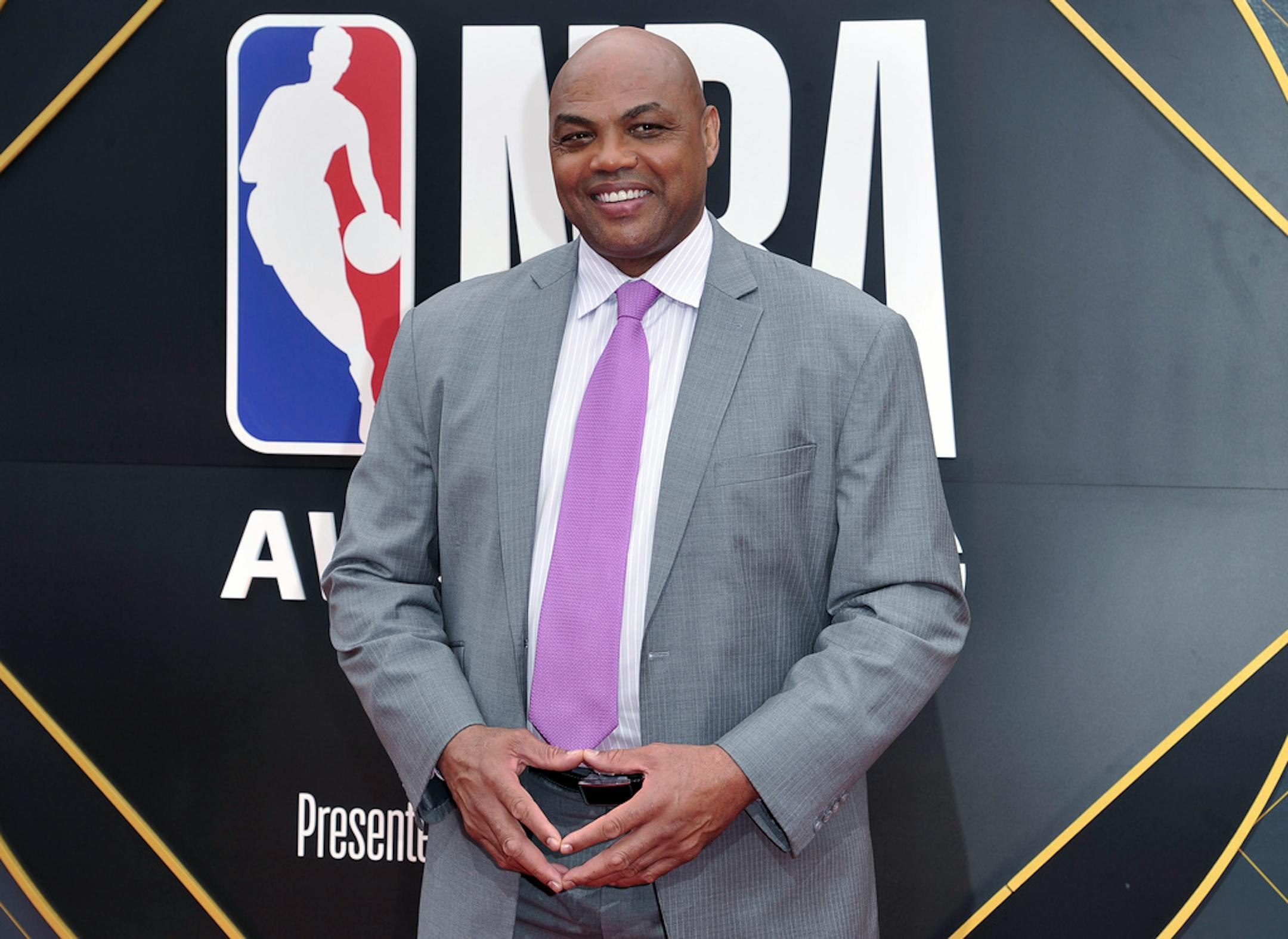 Charles Barkley wearing a gray suit, white shirt and lilac tie in front of an NBA Awards background.