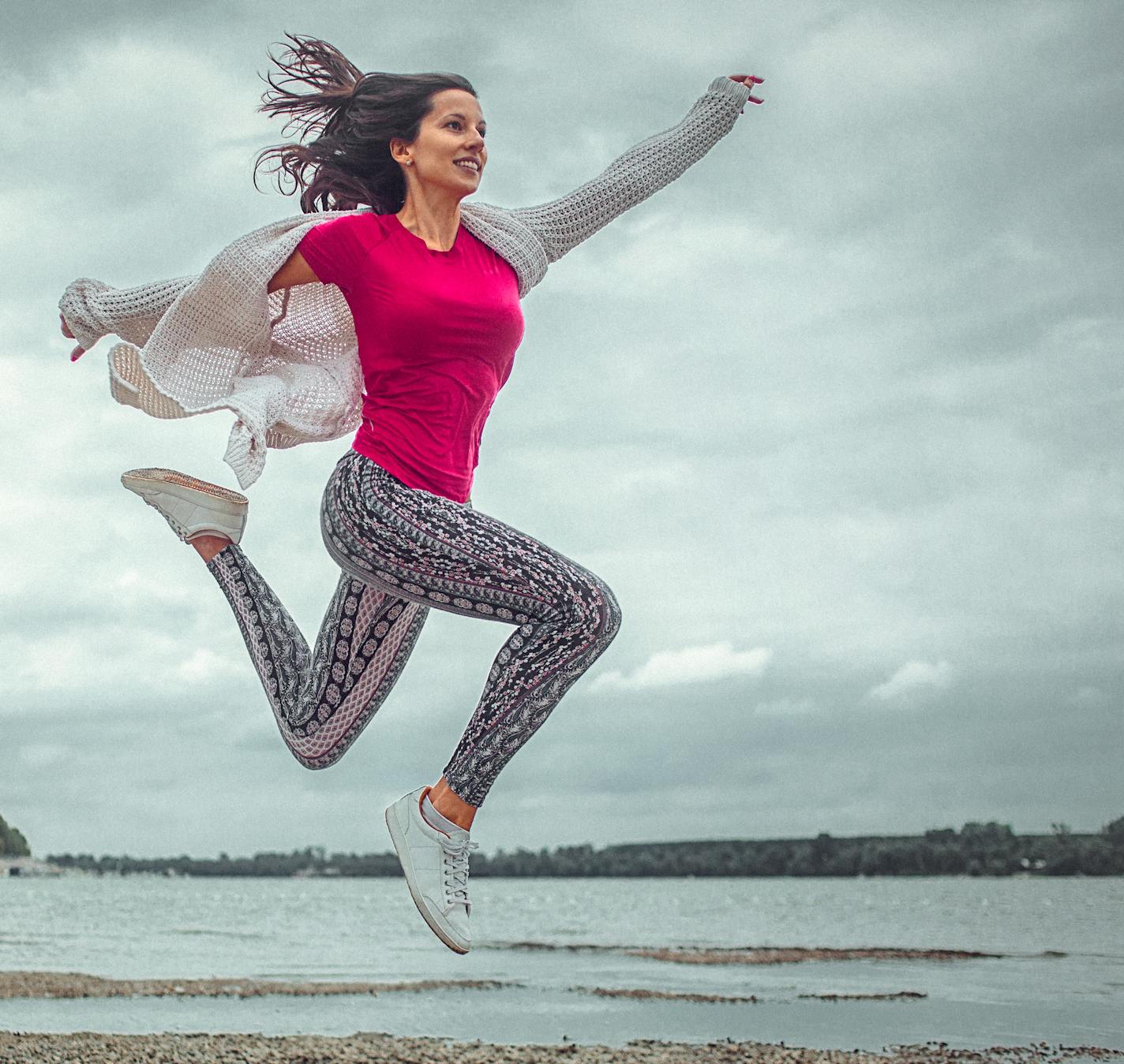 A happy young woman jumping and flying in the air across the beach. The woman's arms and legs are wide spread. She wears red t-shirt, yoga pants and a white vest. The blue cloudy sky is in the background;