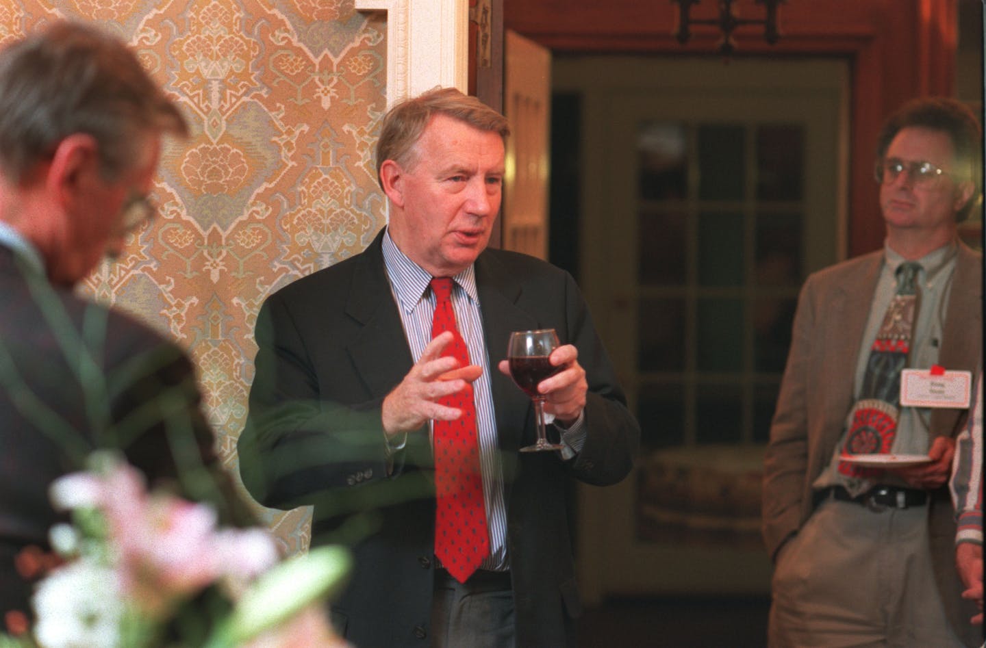 Robert MacNeil wearing a dark sportcoat, striped blue shirt, red foulard tie and gray pants and holding a glass of red wine while talking with a man t