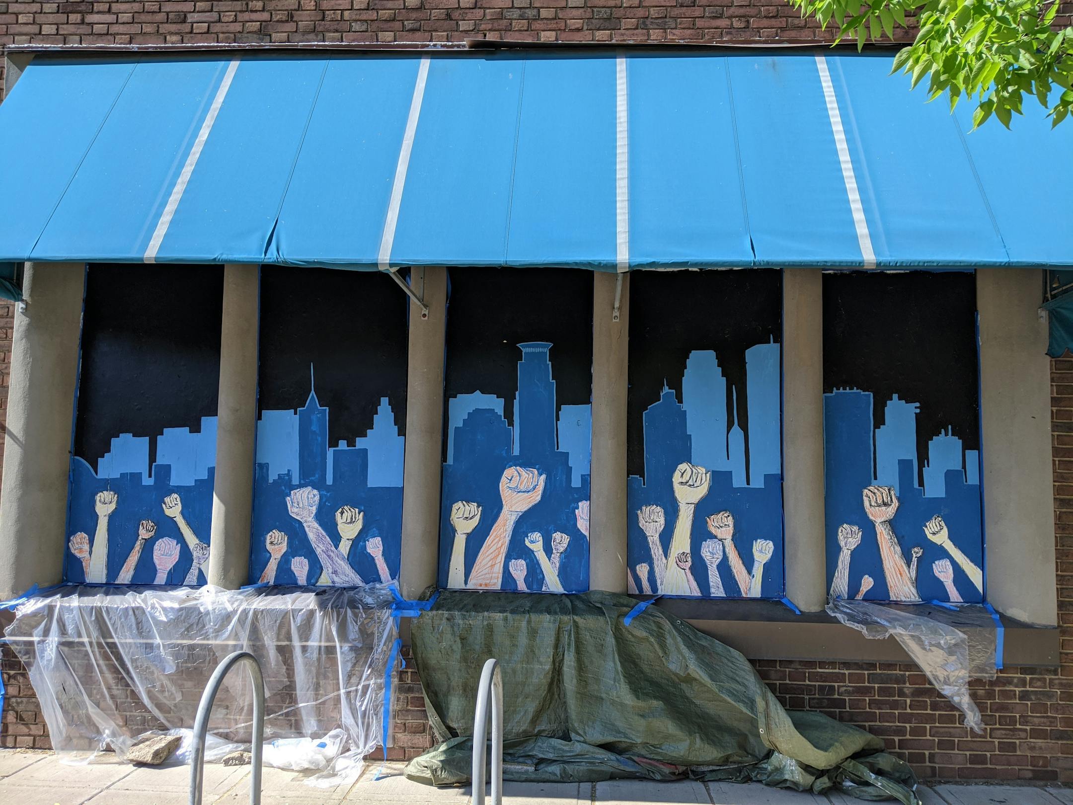 Fists pumping the air in front of a city background cover boarded up windows at Northern Sun at 2916 E Lake St.