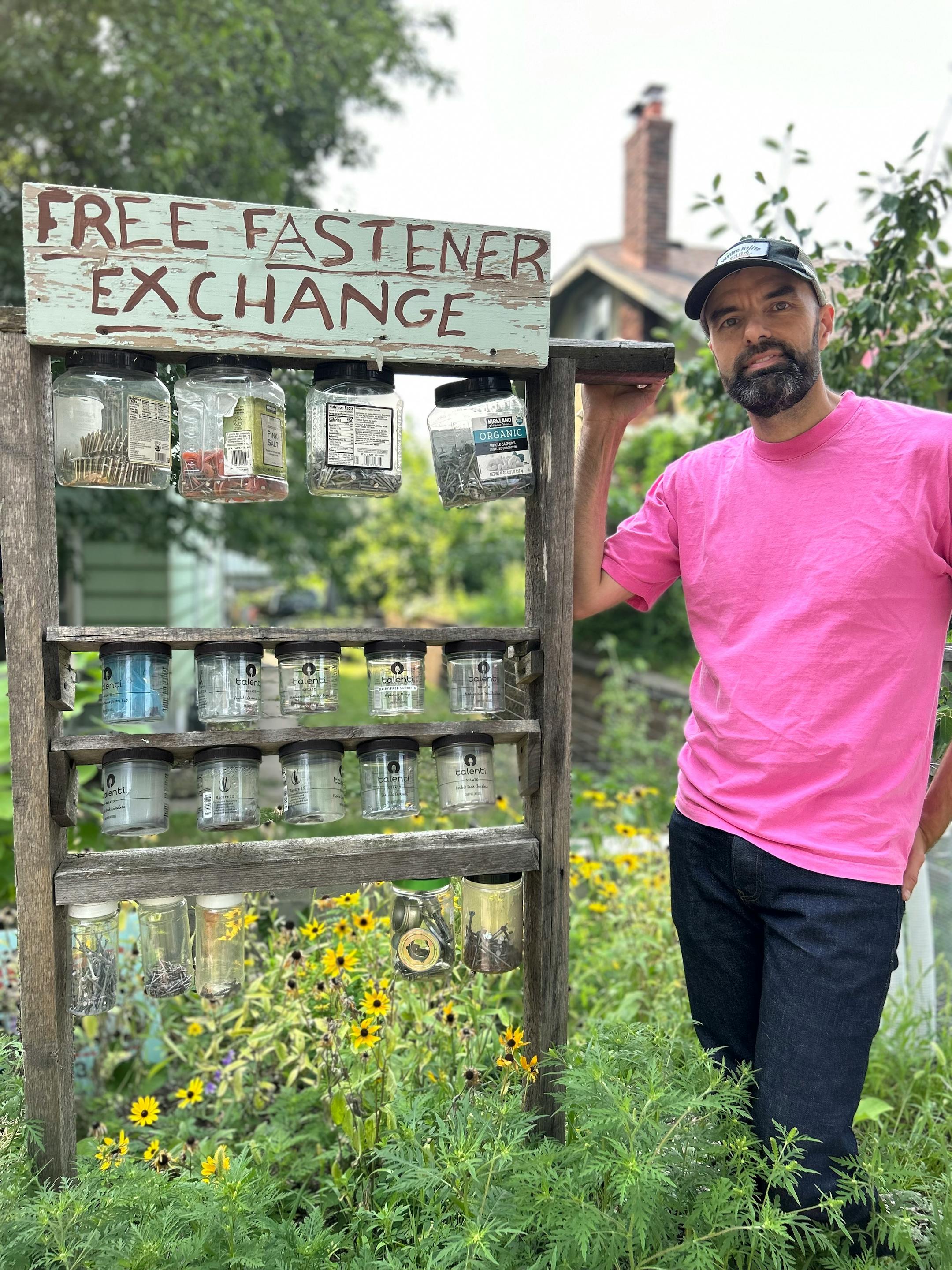 A man wearing a hot pink T-shirt rests his hand on a wooden stand carrying clear plastic jars holding various screws and bolts in a garden filled with thigh-high wildflowers.