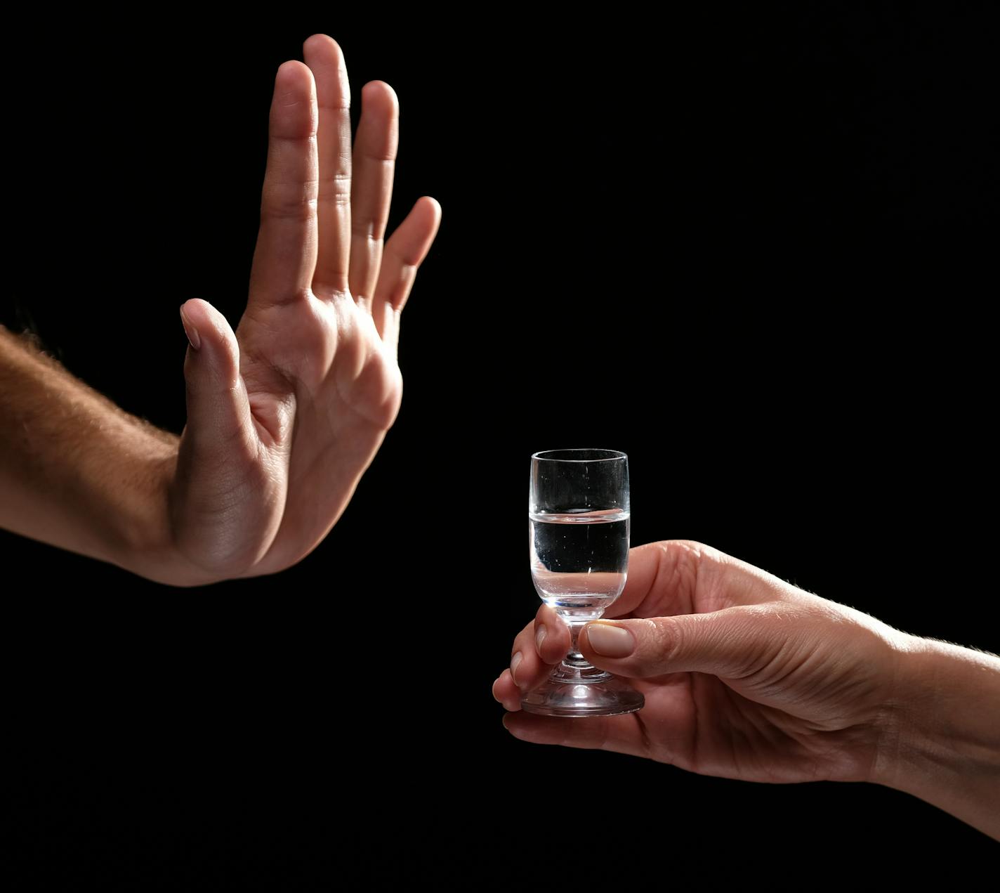 Studio shot black backgroud man's hand in the gesture of refusal vs woman's palm of hand passing glass of alcohol