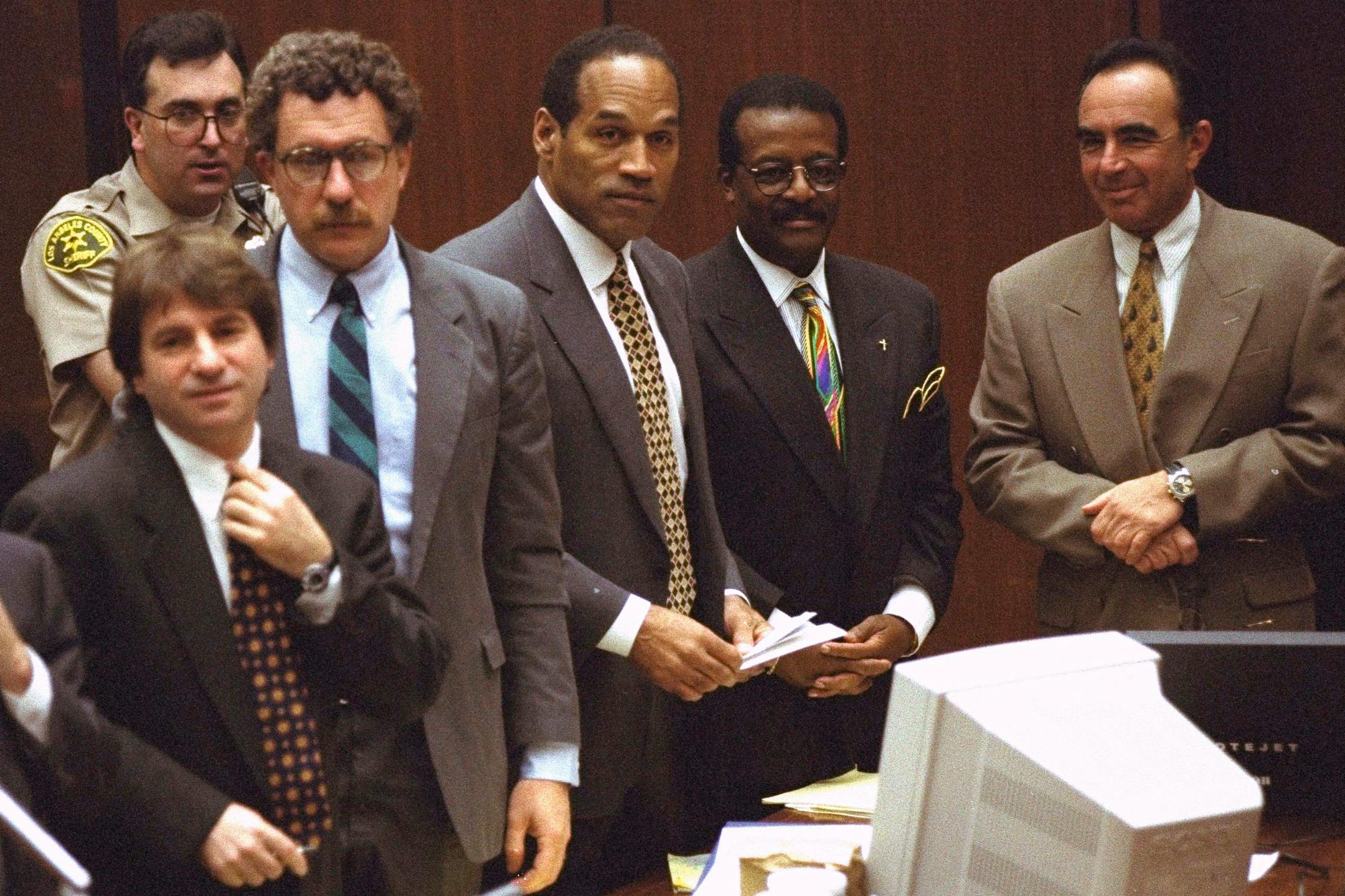 Lawyers and O.J. Simpson in 1990s-style suits standing up with a deputy in the background in the courtroom.