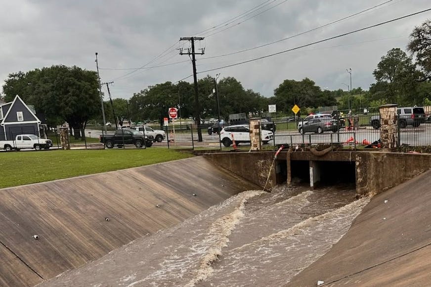 Water flowing through cement ditch with street in background and many vehicles parked.