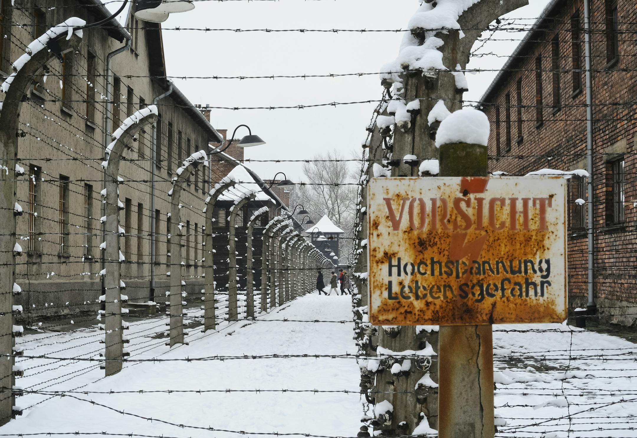 Visitors walk between barbed wire fences at the Auschwitz Nazi death camp in Oswiecim, Poland, Monday, Jan. 26, 2015. A decade ago, 1,500 Holocaust survivors traveled to Auschwitz to mark the 60th anniversary of the death camp's liberation. On Tuesday, for the 70th anniversary, organizers are expecting 300, the youngest in their 70s. Sign in foreground is an electric fence warning.(AP Photo/Alik Keplicz) ORG XMIT: MIN2015012612495356