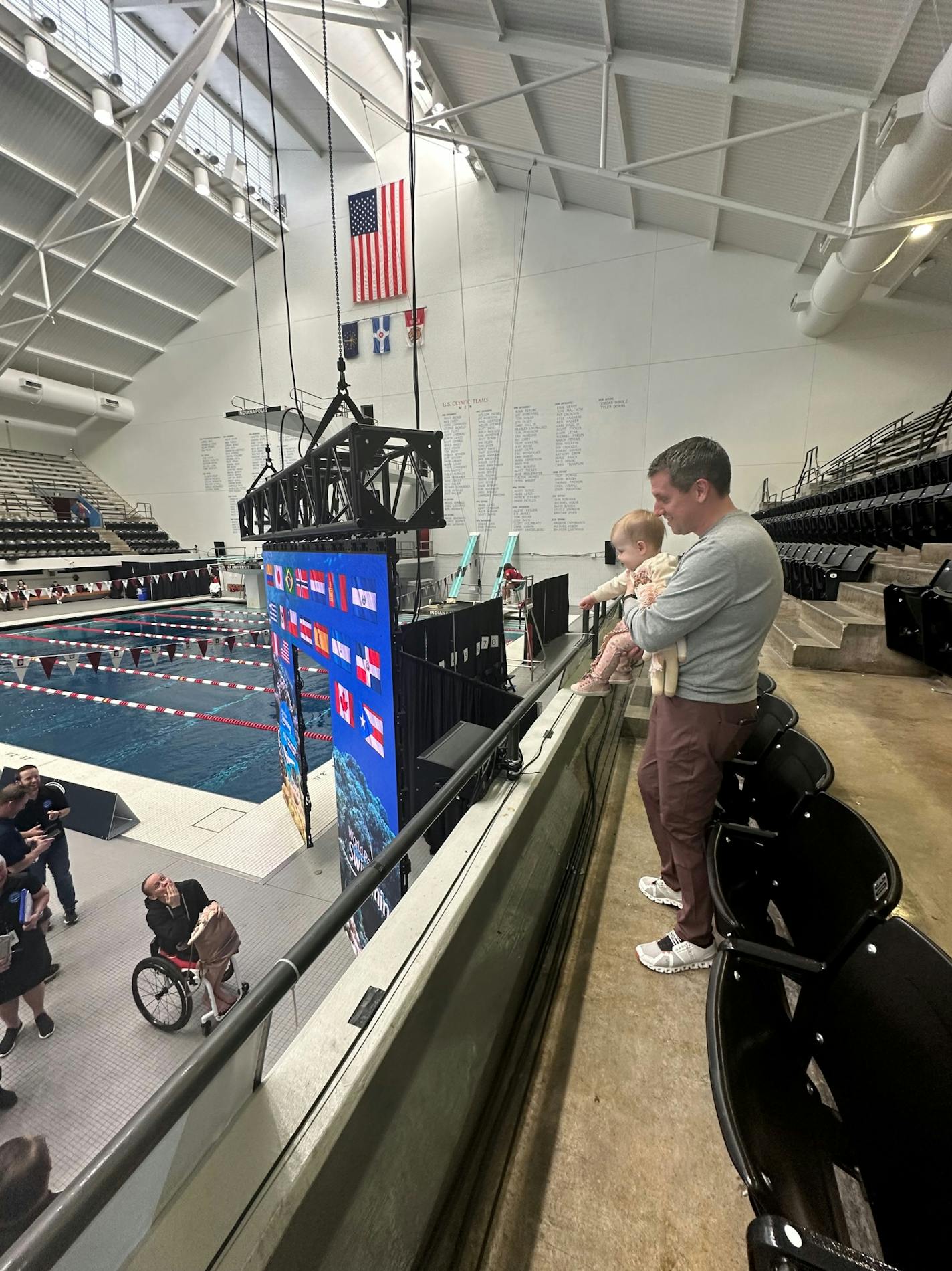 A man holding a baby on the second level of a facility looks over the railing to see his wife in a wheelchair along the pool deck.