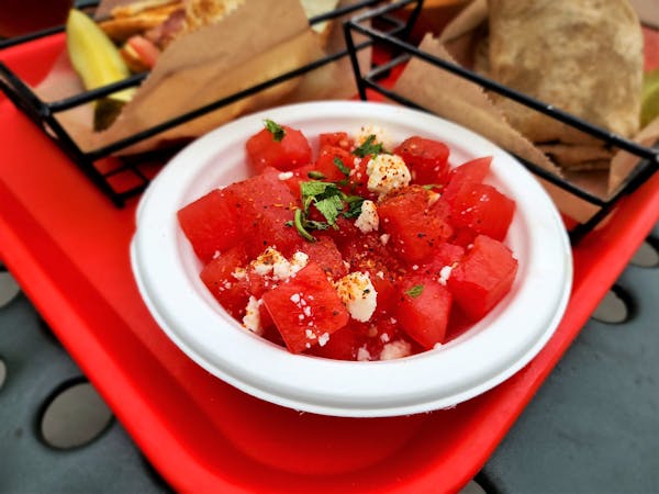 A white bowl filled with cubes of red watermelon sprinkled with a seasoning and some crumbles of white cheese and green herbs, on a red tray.