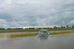 Dark gray or black SUV pointed down into water off road with fields and cloudy sky in the background.