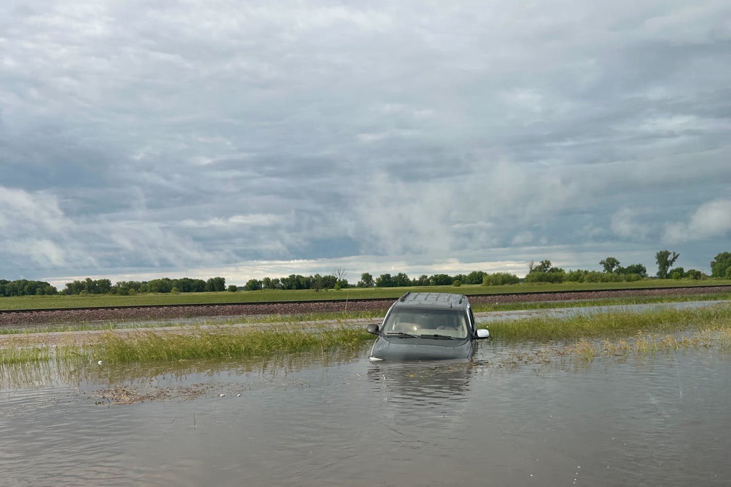 Dark gray or black SUV pointed down into water off road with fields and cloudy sky in the background.