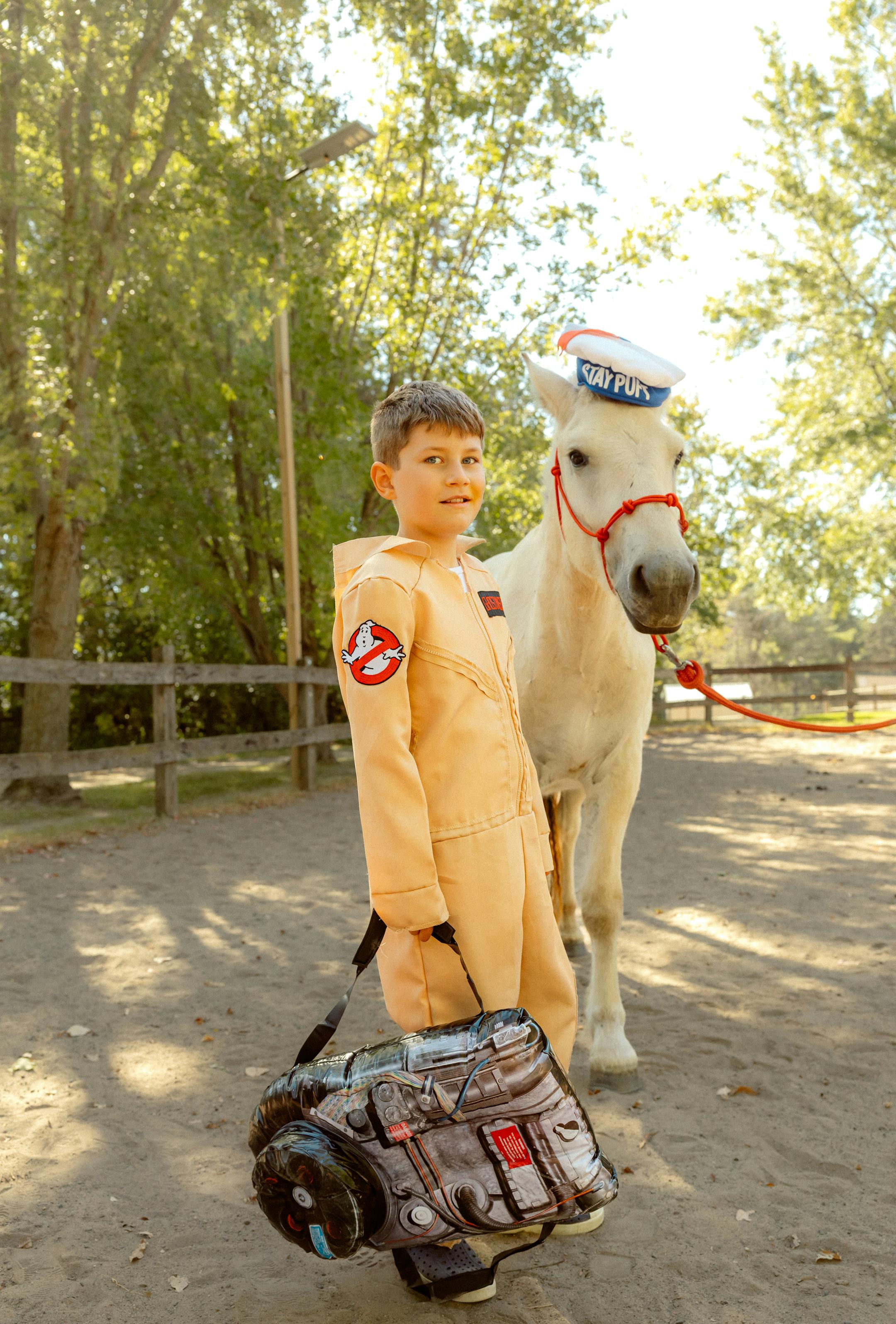 A young teen boy wearing a Ghostbusters coveralls holds a plastic proton-pack while standing next to a white horse wearing a Stay Puft hat.