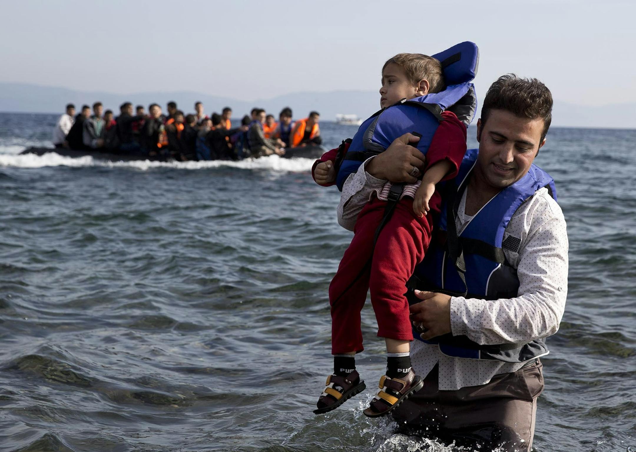 A Syrian refugee carries his child after they arrived from Turkey to the shores of the Greek island of Lesbos, on an inflatable dinghy, with another boat in the background, Saturday Sept. 26, 2015. More than 260,000 asylum-seekers have arrived in Greece so far this year, most reaching the country's eastern islands on flimsy rafts or boats from the nearby Turkish coast. (AP Photo/Petros Giannakouris) ORG XMIT: MIN2015100817561971