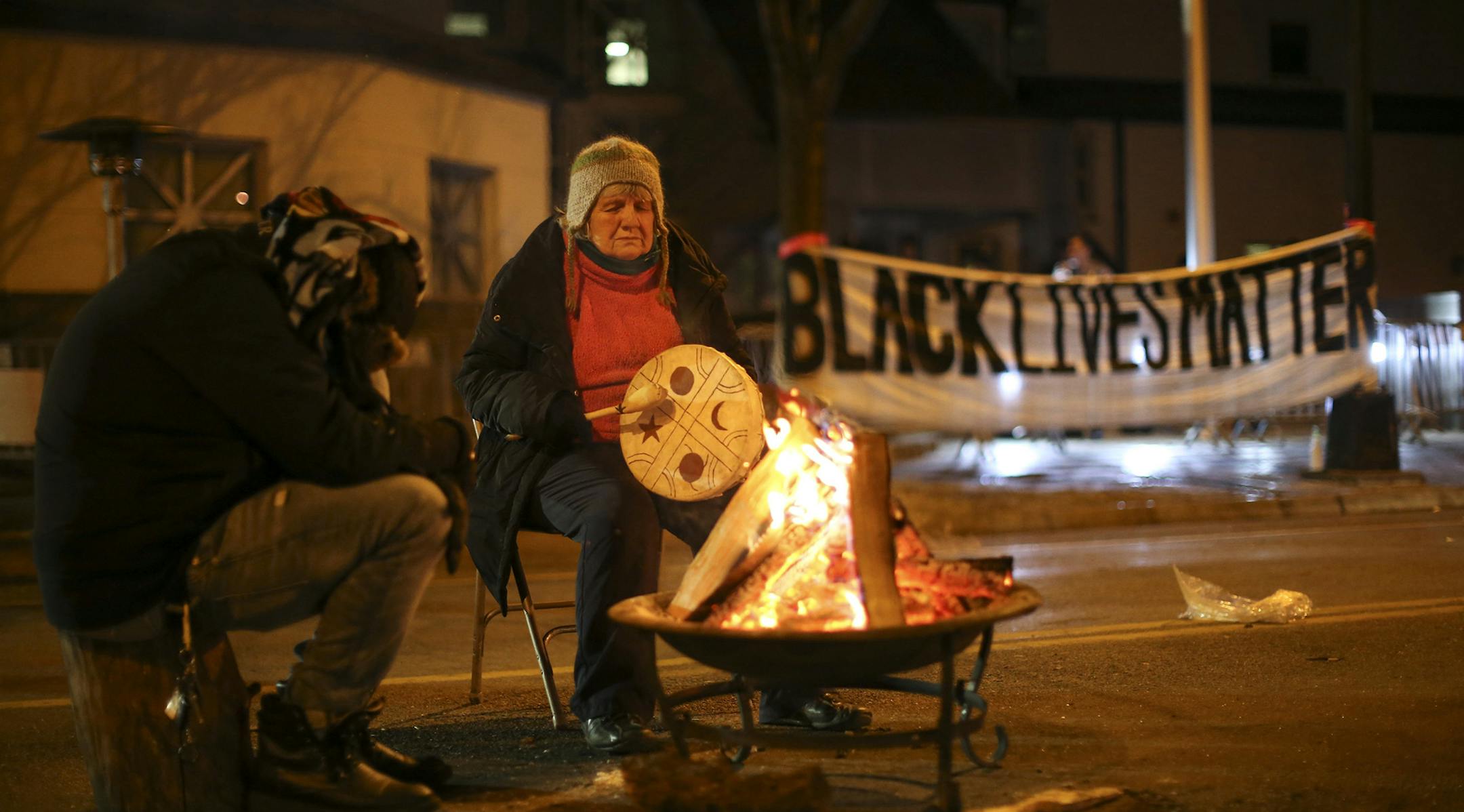 People warm themselves as they demonstrate since the Nov. 10 shooting of 24-year-old Jamar Clark, in front of the Minneapolis Police 4th Precinct on Tuesday, Nov. 24, 2015. Minneapolis police were searching Tuesday for three white males suspected of shooting at five Black Lives Matter demonstrators, while the family of a black man who was fatally shot by a city police officer called for the dayslong protests outside of the police precinct to end. (Jeff Wheeler/Star Tribune via AP) ORG XMIT: MIN2