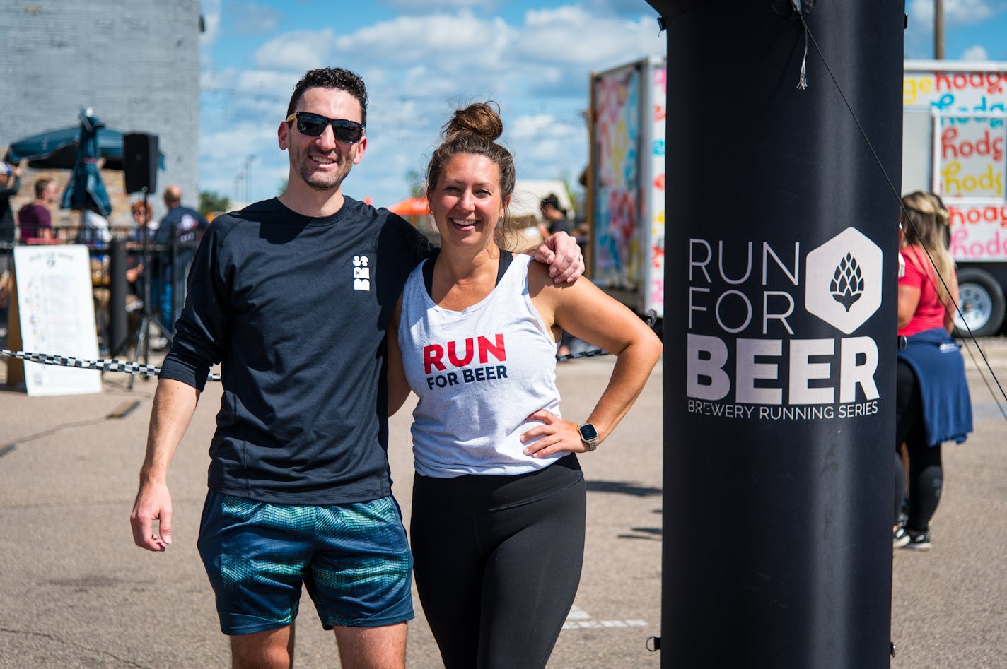A man with sunglasses and running shorts/shirt puts his arm around the shoulder of a woman with a tank top and leggings in front of an inflatable arch that says "Run for Beer."
