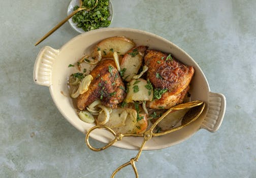 A cream oval baking dish with chicken thighs, apples and fennel sprinkled with fresh herbs.