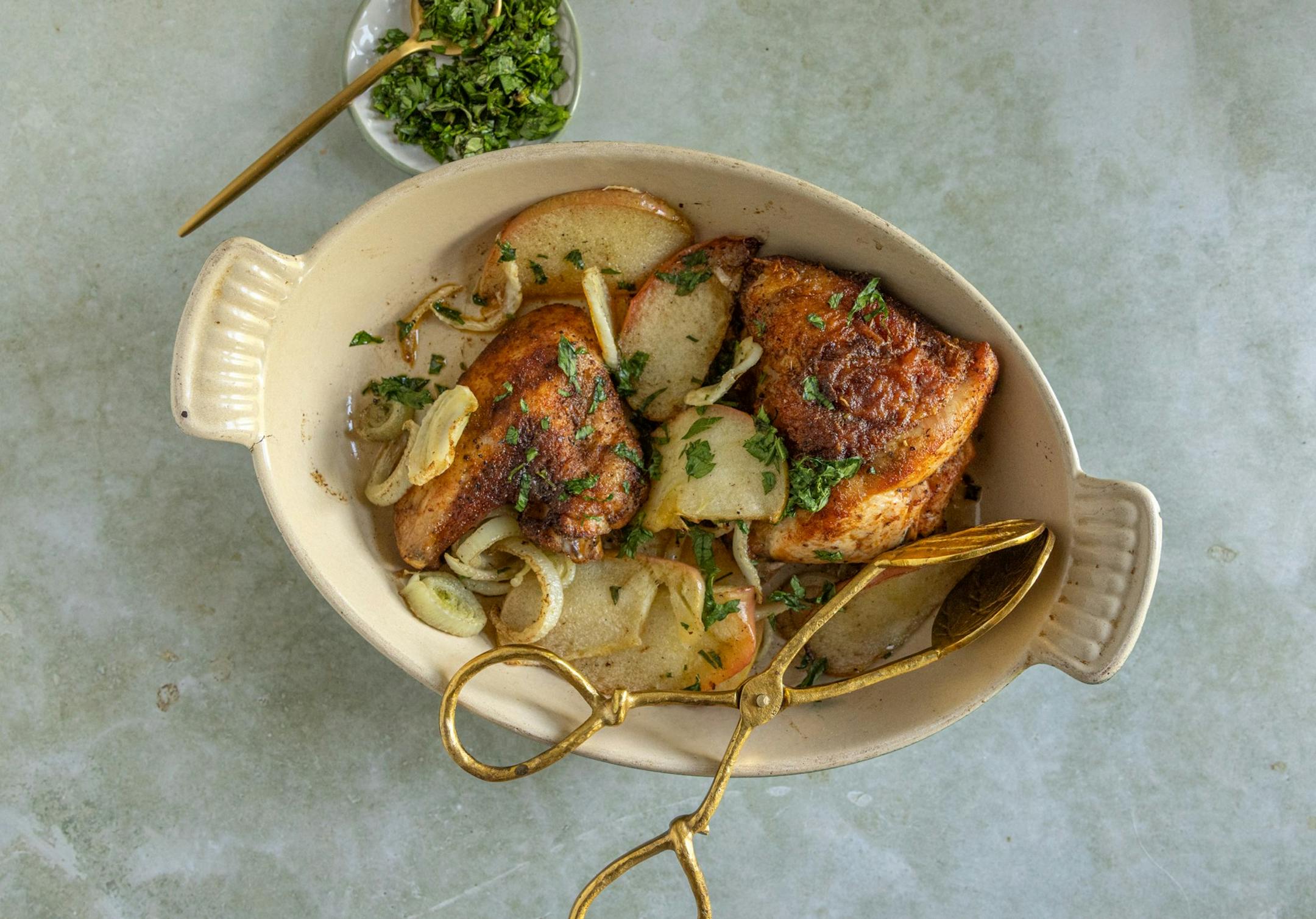 A cream oval baking dish with chicken thighs, apples and fennel sprinkled with fresh herbs.