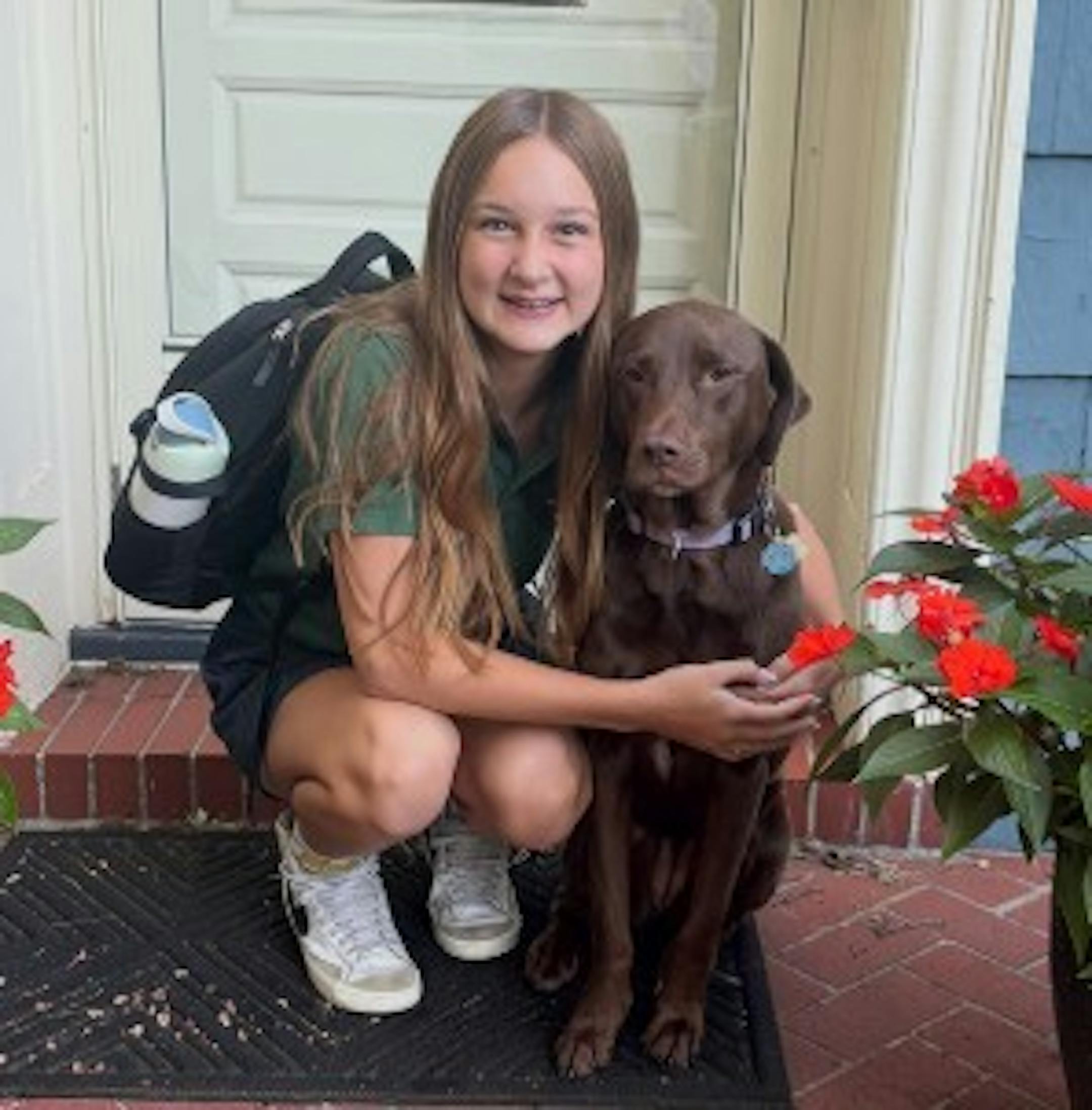 A 12-year-old girl with a backpack and green polo school uniform shirt poses with her arms around a chocolate Lab on a front stoop of a home.