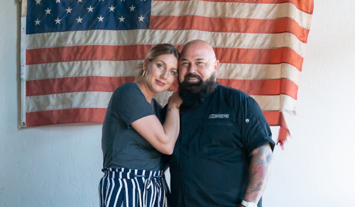 Sarah leans on Brian's shoulder. He's wearing a black chef's coat in front of a large American flag inside their original Hope Breakfast Bar in St. Paul