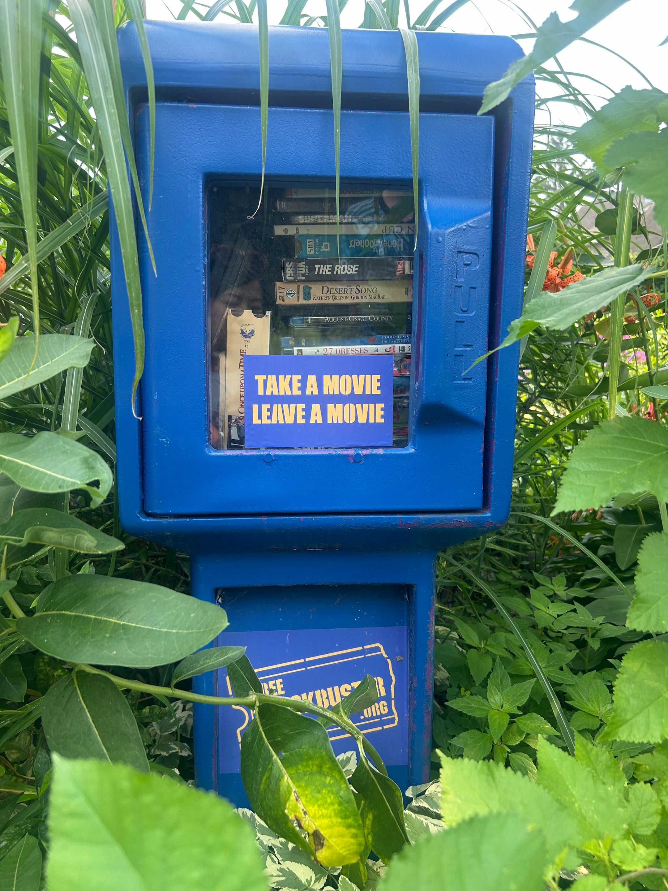 A blue and yellow former newspaper rack contains a big stack of DVDs and VHS tapes.