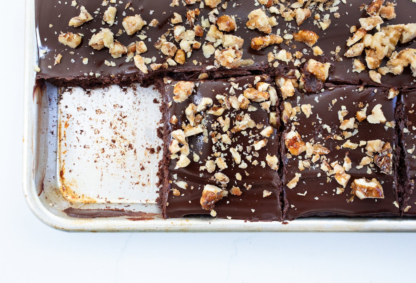 A sheet pan of chocolate cake topped with chocolate ganache and candied black walnuts with one piece missing.