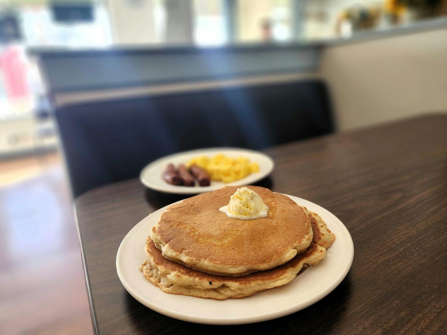 On a diner table, a plate with two large pancakes and a melting scoop of butter. In the background, a plate with scrambled eggs and sausage.