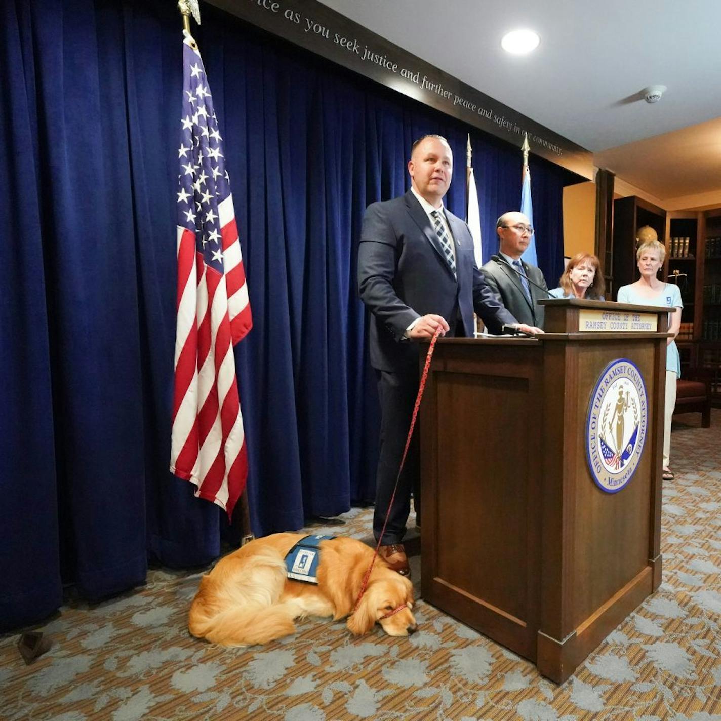 Norie, the new RCAO Facility Dog came into the press conference with her handler, Bill Kubes, RCAO Victim Advocate, and stood or laid by his side as cameras recorder her every move. L to R are Norie, RCAO Facility Dog, Bill Kubes, RCAO Victim Advocate, Norie's handler, John Choi, Ramsey County Attorney, Tami McConkey, RCAO Director of Victim/Witness Division Sue Kliewer, Helping Paws Client Services Coordinator and Instructor.