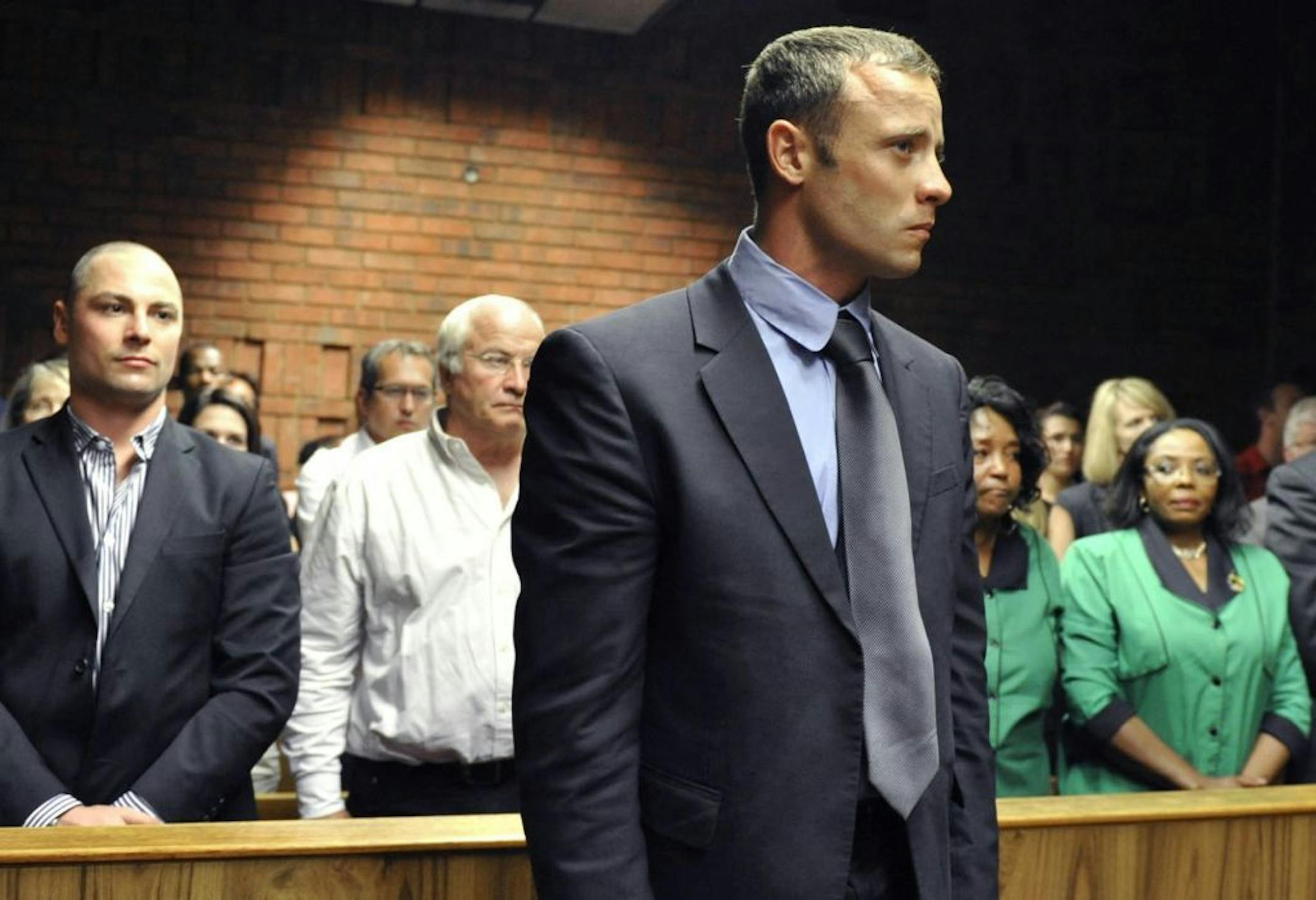 Olympian Oscar Pistorius, foreground, stands following his bail hearing, as his brother Carl, left, and father Henke, second from left, look on in Pretoria, South Africa, Tuesday, Feb. 19, 2013. Pistorius fired into the door of a small bathroom where his girlfriend was cowering after a shouting match on Valentine's Day, hitting her three times, a South African prosecutor said Tuesday as he charged the sports icon with premeditated murder. The magistrate ruled that Pistorius faces the harshest ba