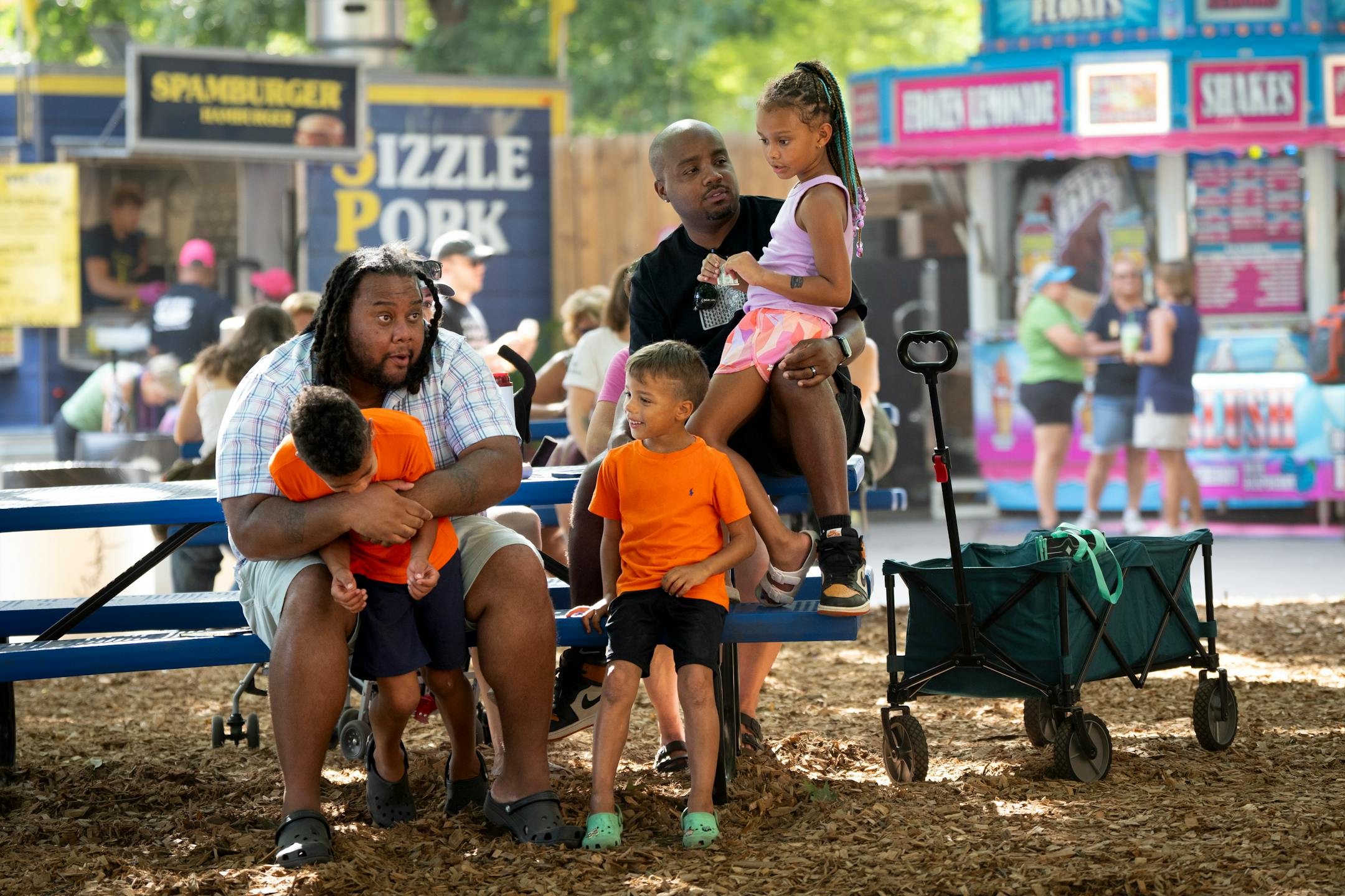 Switching kids with his friend, Torrean Davison tackle hugs Kingston Stong, 8, as Darwin Strong hold's Davison's daughter Rainy Davison on his lap while chilling in the shade on the first day of the Minnesota State Fair on Thursday, Aug. 24, 2023 in Falcon Heights, Minn. Also pictured is Kayson Strong, 5.