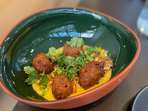 Four fried corn fritters in a pool of bright yellow sauce in a large bowl/plate that's a forest green enamel on the inside.