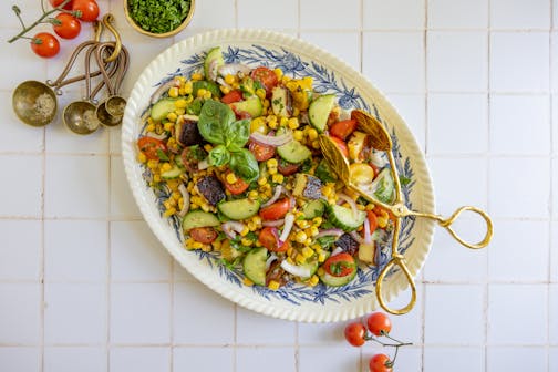 A large platter with a salad of cherry tomatoes, cucumbers, sweet corn, bread cheese and fresh herbs.