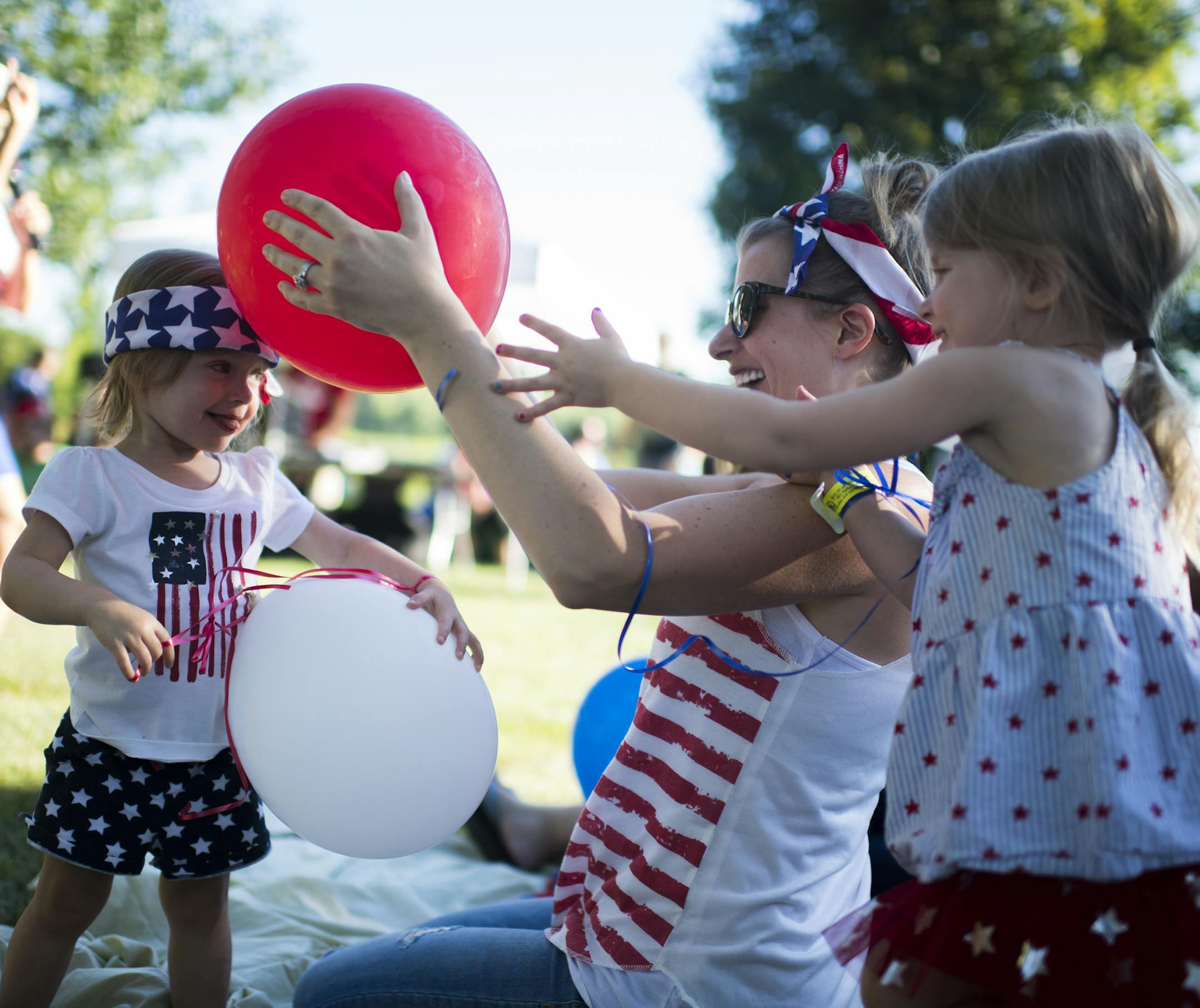 Reagan Weber, 2, smiled as her mom, Kristine, and sister, Ellyson, 3, created static electricity with a balloon on her head as they listen to a children's band play before the fireworks display later in the evening.