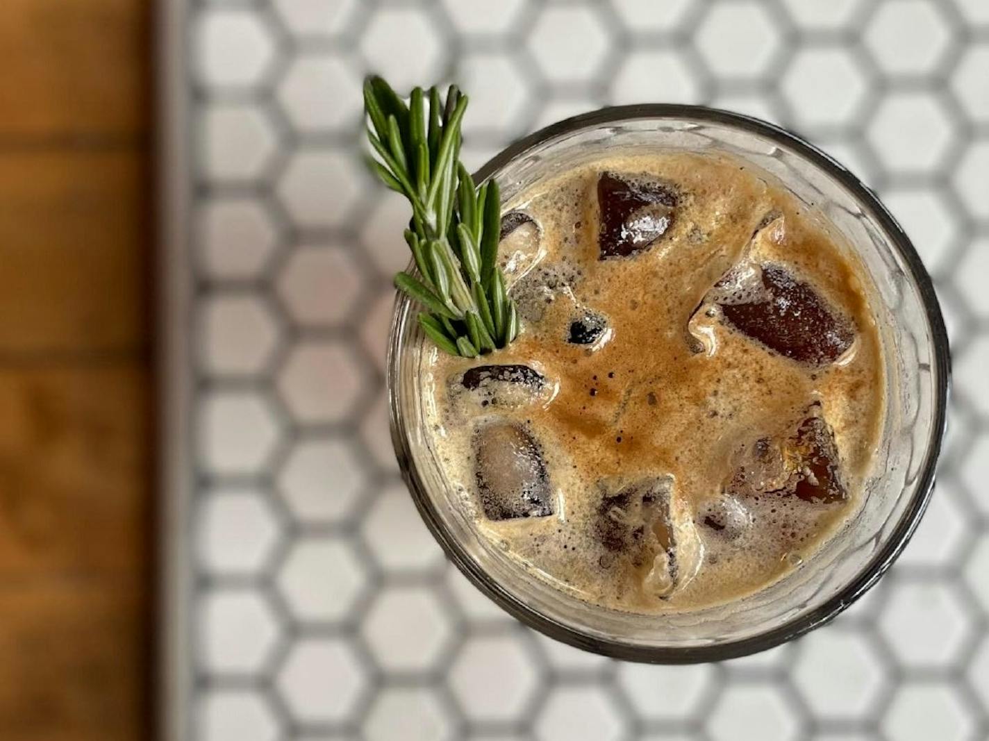 A pint glass against black and white penny tile is filled with a fizzy coffee drink, garnished with rosemary.