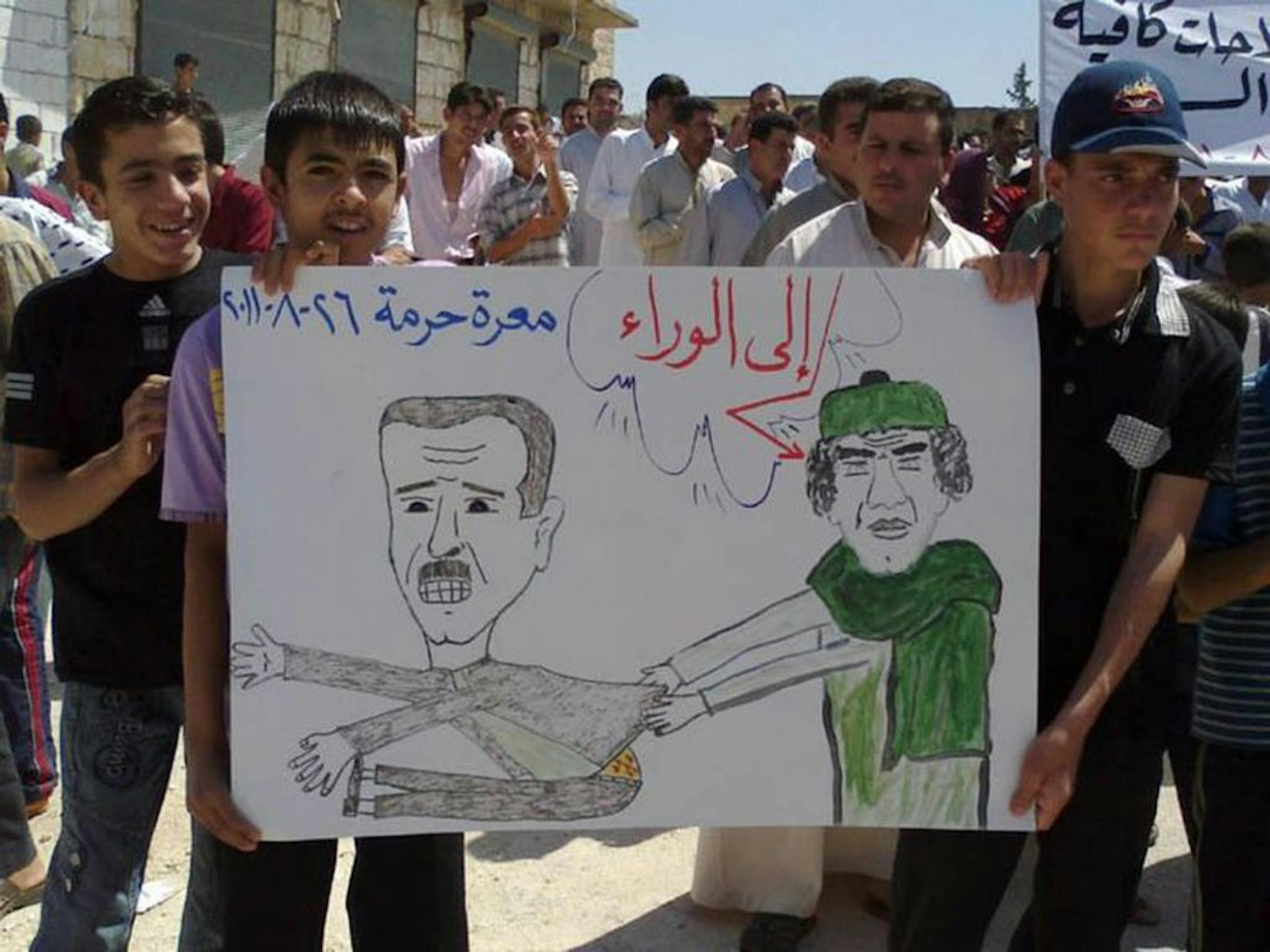 In this citizen journalism image made on a mobile phone and provided by Shaam News Network, anti-Syrian President Bashar Assad protesters, hold a cartoon placard depicting Moammar Gadhafi, right, and Syrian President Bashar Assad, left, with Arabic words read:"step back," during a demonstration against the Syrian regime, at Maaret Harma village, in Edlib province, Syria, on Friday Aug. 26, 2011. Syrian security forces killed at least two people as tens of thousands of anti-government protesters