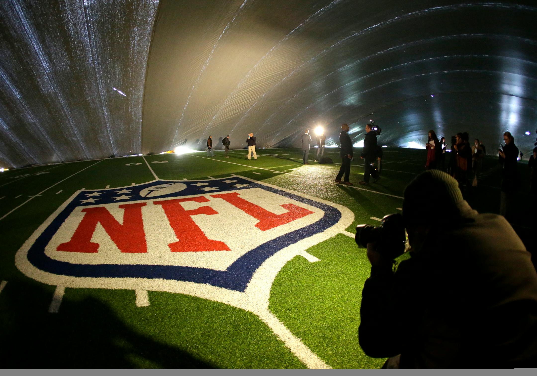 FILE - In this Jan. 15, 2014 file photo, the NFL logo at midfield of MetLife Stadium is illuminated by lights on television reporters' videocameras as members of the media are given a tour under a tarp used by crews to keep the turf dry ahead of Super Bowl XLVIII in East Rutherford, N.J. As the Denver Broncos and Seattle Seahawks prep for their Feb. 2, 2014 showdown in the Super Bowl, a legal fight is playing out over the very turf installed months ago at the NFL title game's venue. Taylor Turf