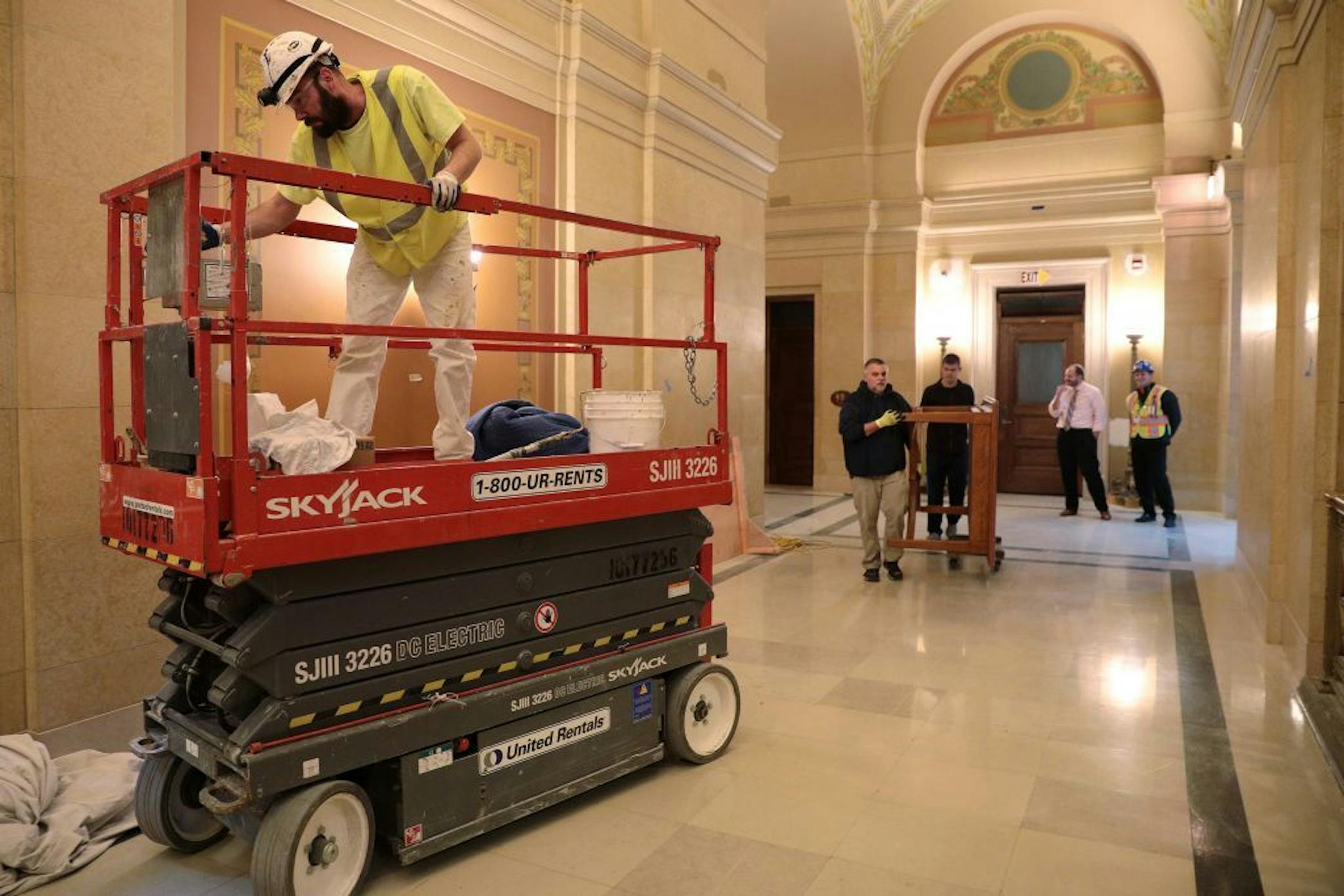 Historical painter Edward Peterson moves his scissor lift so workers can move a table down the hallway at the capital rotunda's east side Wednesday.