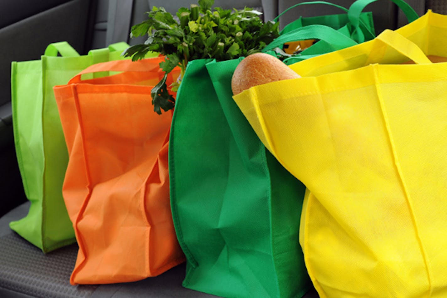 Four colorful eco-friendly shopping bags filled mostly with groceries in the back seat of a car.