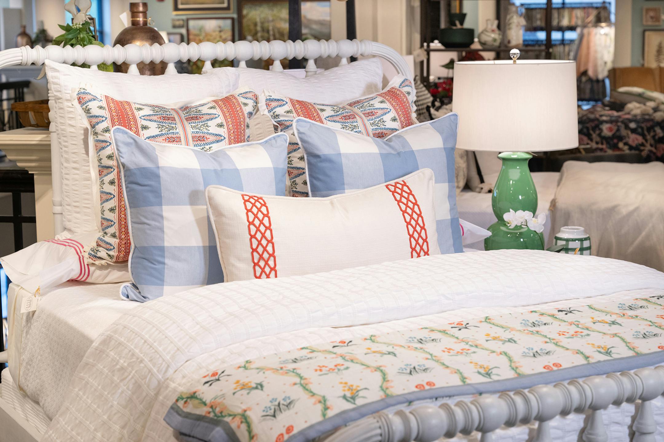 A white spindle bed is layered with pillows, starting with a bolster in front and then three layers of two square pillows. White, coral and pale blue are the main colors.